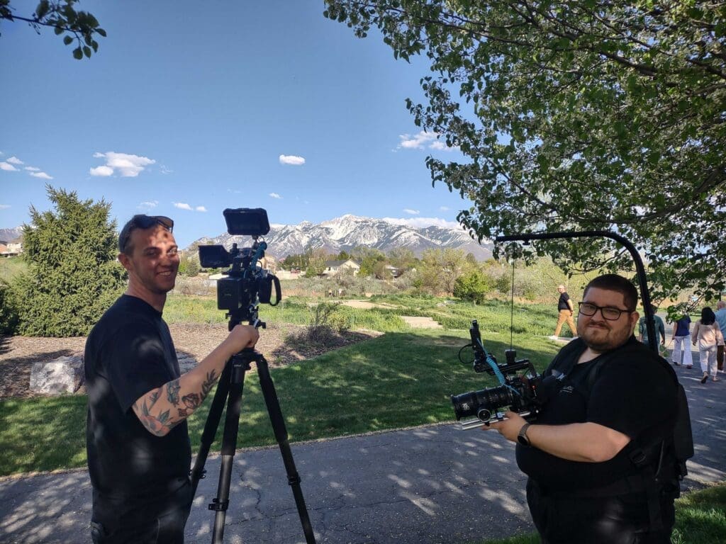 Two members of a film crew outdoors, both managing camera equipment, with mountains under a clear sky in the background.