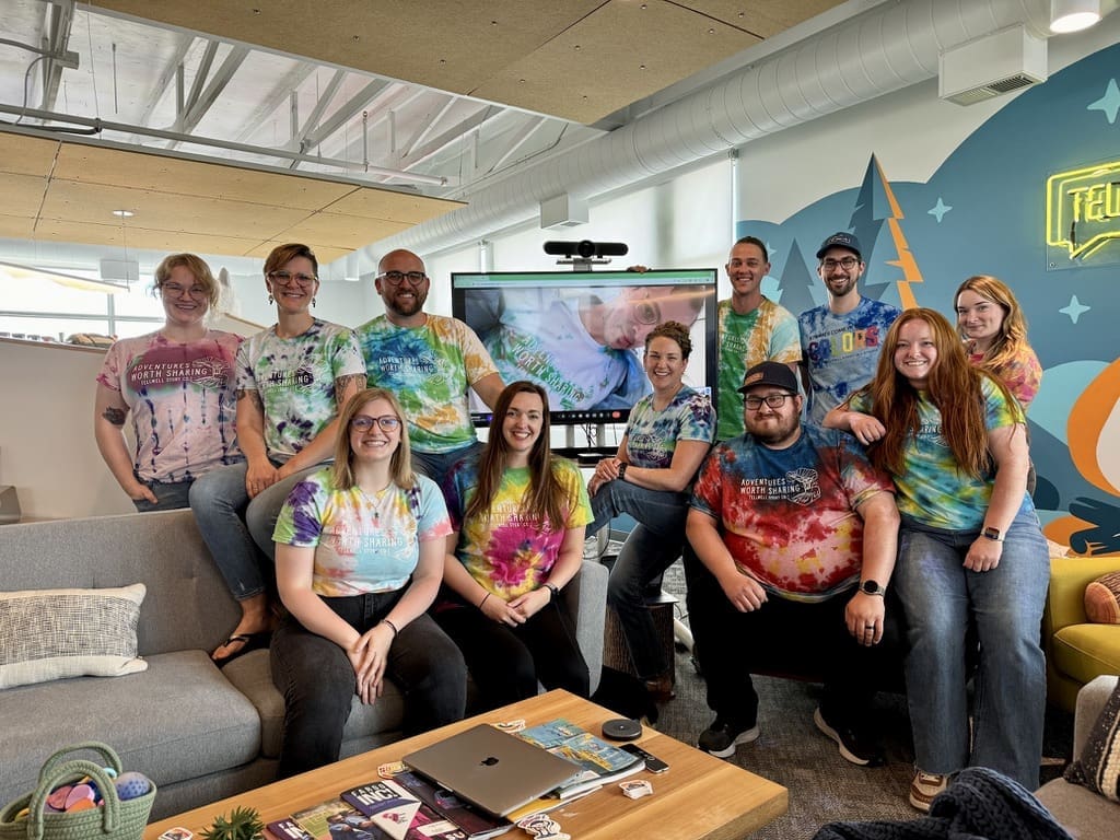 A group of eleven people are gathered together in a brightly lit office space. They are all wearing colorful tie-dye T-shirts that read "Adventures Worth Sharing." Some are seated on a gray couch while others stand behind it. A TV screen in the background displays an image of one of the group members who is participating via video call. The office features modern decor, including a wall mural with trees and a neon sign that says "Tellwell." The atmosphere appears cheerful and collaborative.