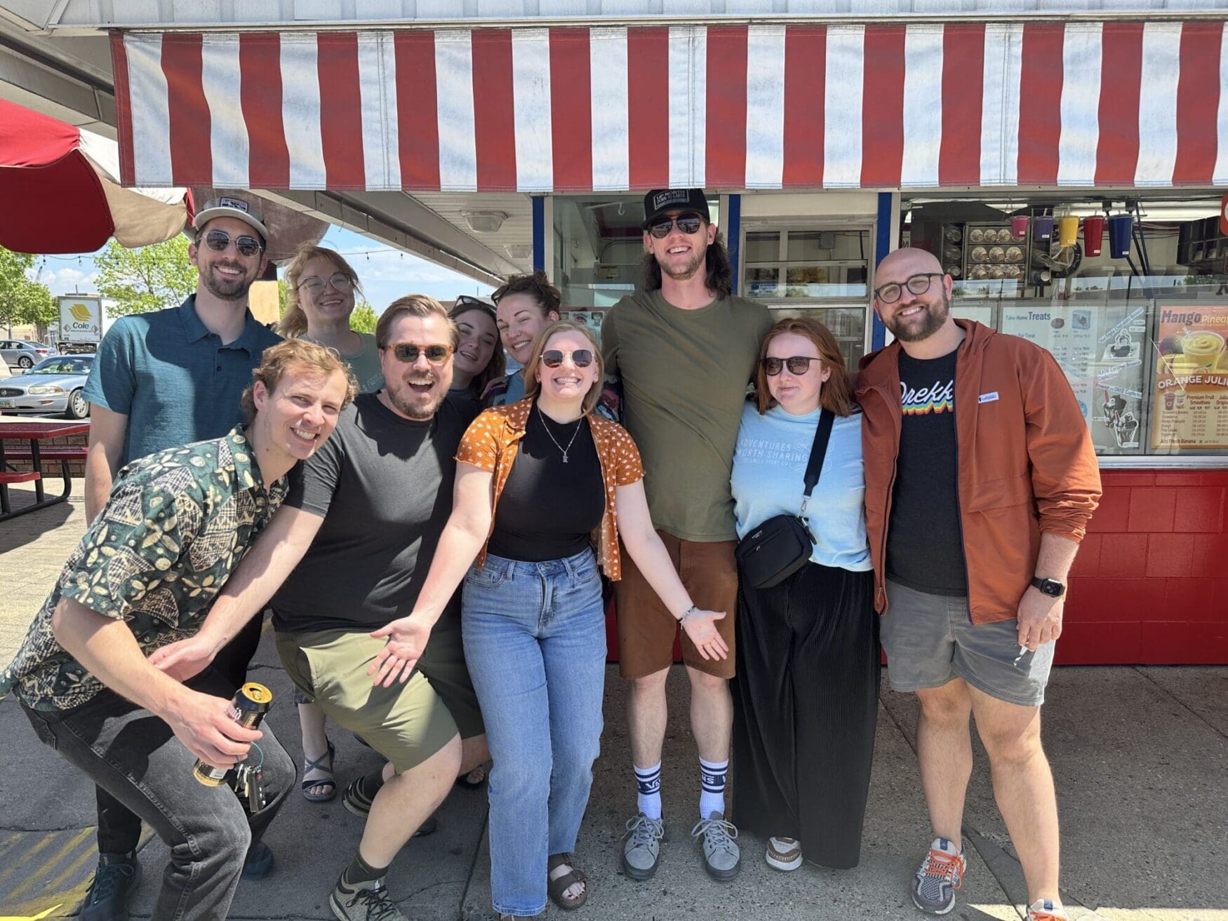 A group of nine people is standing in front of a red and white striped awning, smiling and posing for a group photo. They are outside a food establishment.