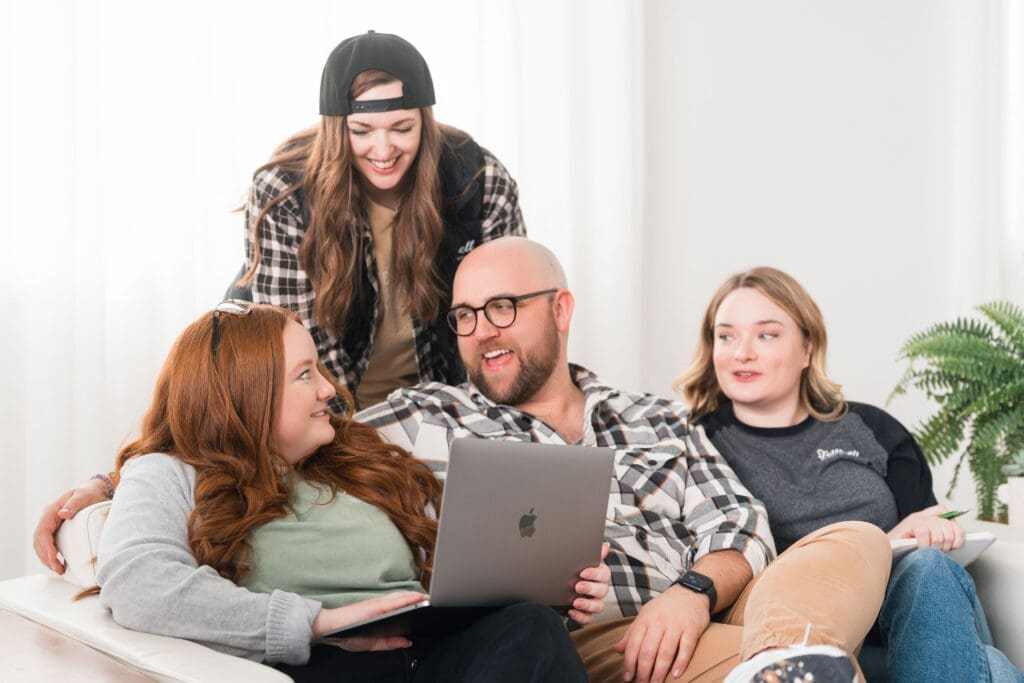 Four people are sitting and interacting on a white couch. A man with a beard and glasses holds a laptop, smiling and talking to a red-haired woman next to him. Another woman with long hair and a backwards baseball cap is standing behind them, smiling and looking down at the group. A fourth person with shoulder-length blonde hair is seated at the end of the couch, holding a notebook. A potted plant is visible in the background.