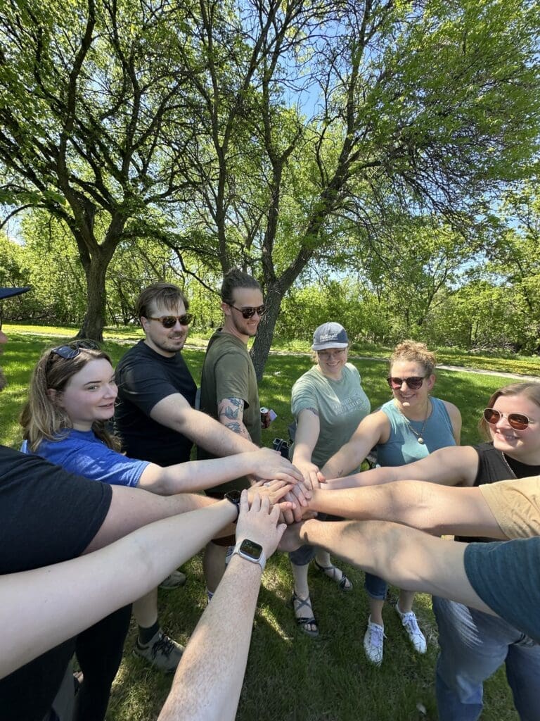 A group of people standing in a circle outdoors, placing their hands together in the center as a gesture of unity. They are in a park with green grass and large trees, under a bright blue sky. Everyone is smiling and enjoying the moment.