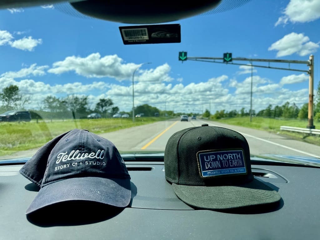 Two hats are displayed on the dashboard of a car. The hat on the left is a navy blue baseball cap with the words 'Tellwell Story Co. & Studio' embroidered in white. The hat on the right is a black snapback cap with a patch that reads 'UP NORTH DOWN TO EARTH' and 'Tellwell Story Co. & Studio' underneath. The view through the windshield shows a sunny day with a blue sky, scattered clouds, and a road with green fields and trees on either side.