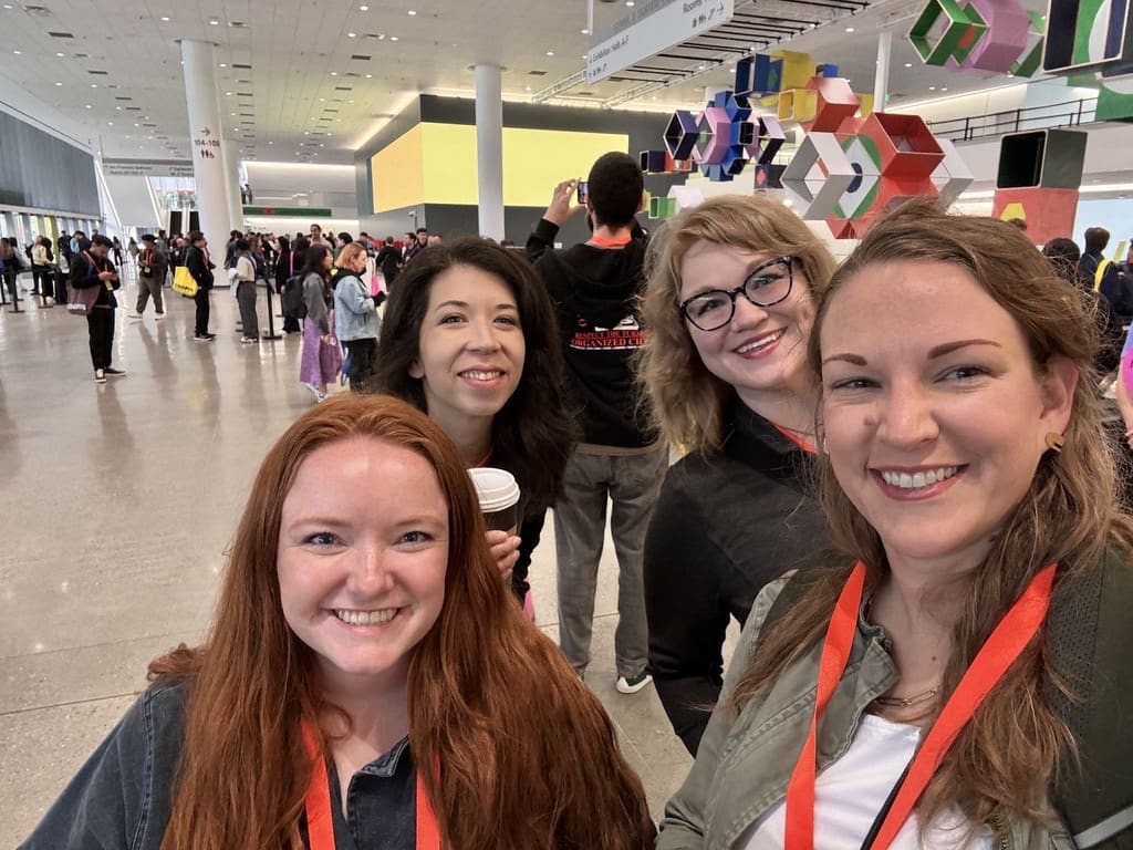 Four smiling women with conference badges take a selfie in a large, busy convention center. The background shows a crowd of attendees, modern decor with geometric shapes, and large digital screens. The women appear happy and excited to be at the event.