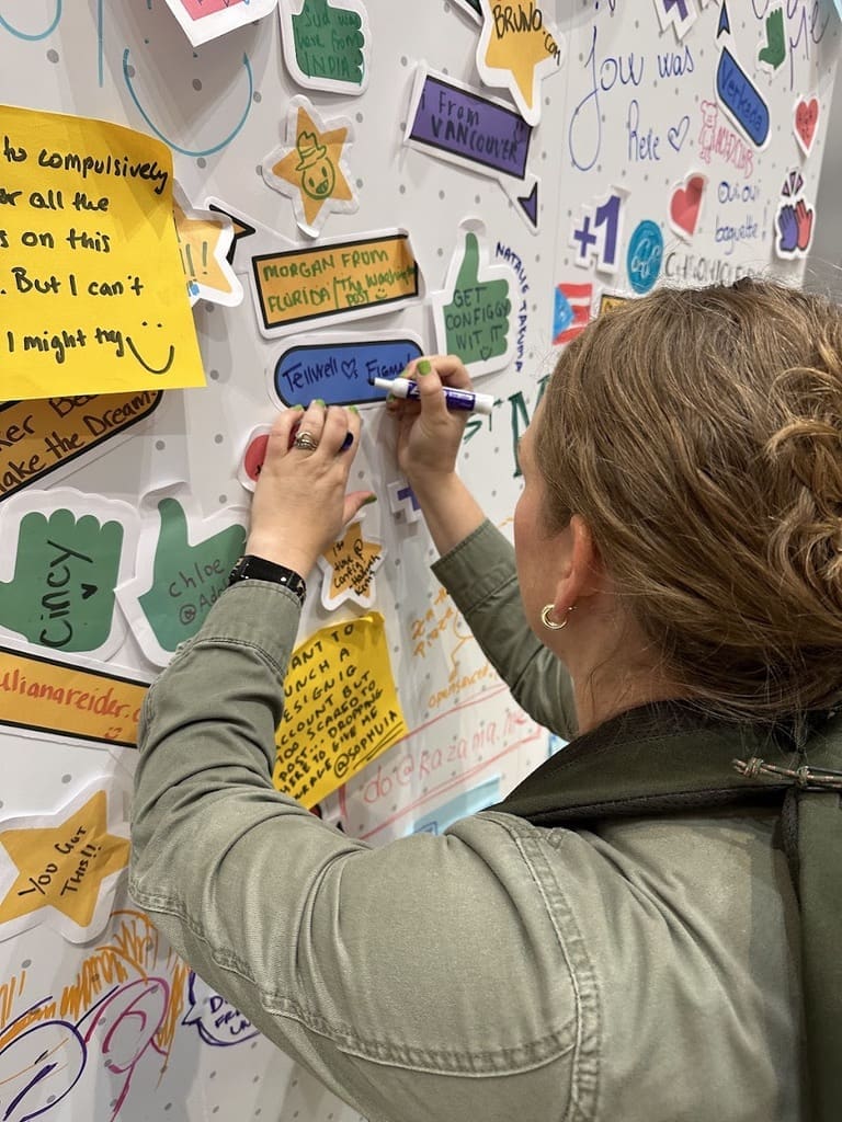 A woman writing on a colorful wall filled with sticky notes and messages at a conference. She is using a blue marker to add her note, surrounded by various handwritten messages and drawings. The atmosphere appears creative and engaging, with contributions from multiple participants.