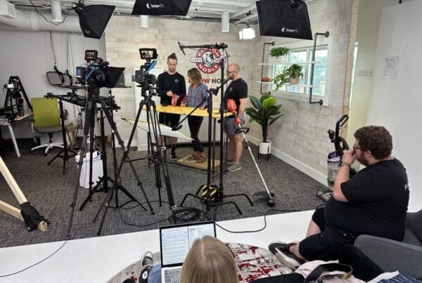A behind-the-scenes view of a filming studio setup with multiple cameras, lights, and crew members. In the foreground, a person is sitting on a couch with a laptop. In the middle, three people are standing around a table discussing or preparing something. The studio has a modern and organized setup with various equipment and plants decorating the space.