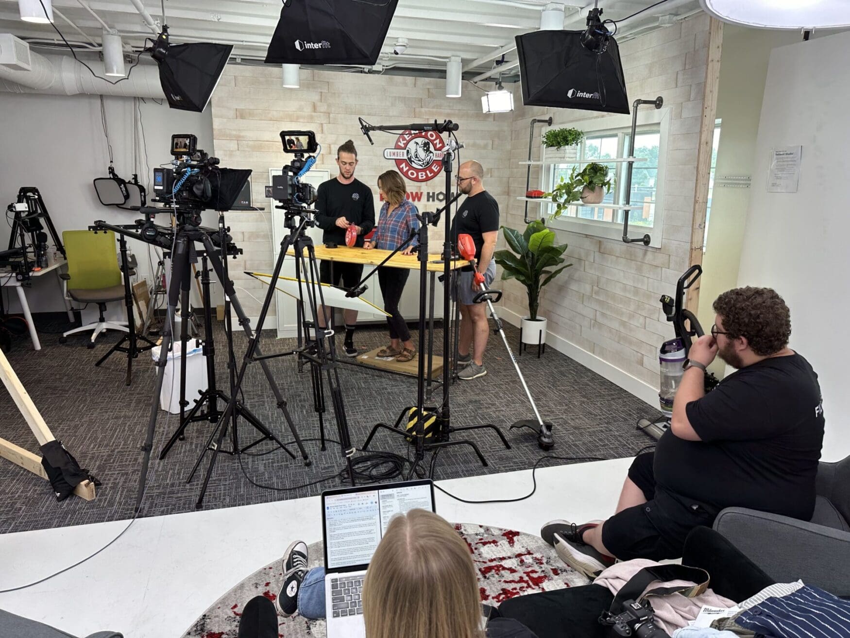 A behind-the-scenes view of a filming studio setup with multiple cameras, lights, and crew members. In the foreground, a person is sitting on a couch with a laptop. In the middle, three people are standing around a table discussing or preparing something. The studio has a modern and organized setup with various equipment and plants decorating the space.