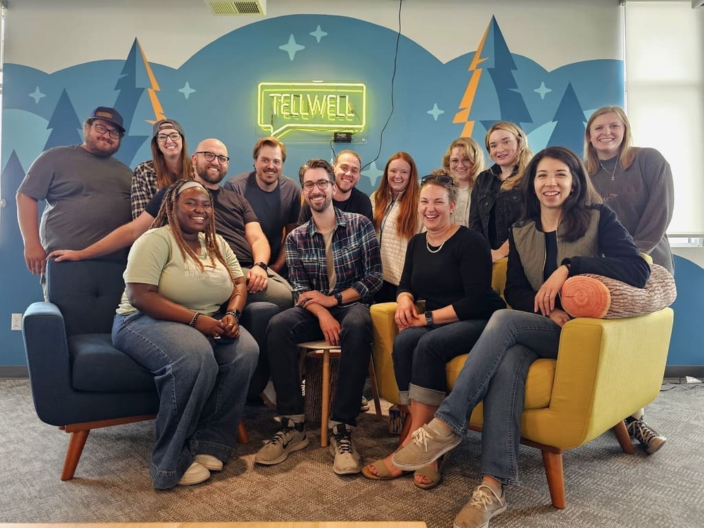 A group of 13 people, smiling and casually dressed, are gathered in an office space with a bright and cheerful atmosphere. They are sitting and standing around two modern armchairs, with a neon "TELLWELL" sign on the wall behind them. The background features a blue mural with mountain and tree designs, creating a cozy and creative environment. The group appears to be close-knit and happy, reflecting a positive team spirit.
