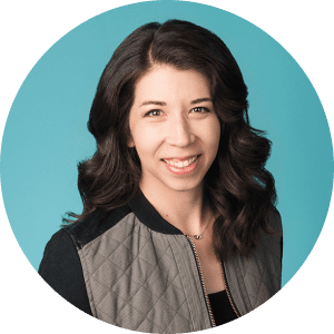 Portrait of Jennifer Klath, a designer, smiling with wavy dark hair, wearing a quilted jacket and a delicate necklace against a teal background.