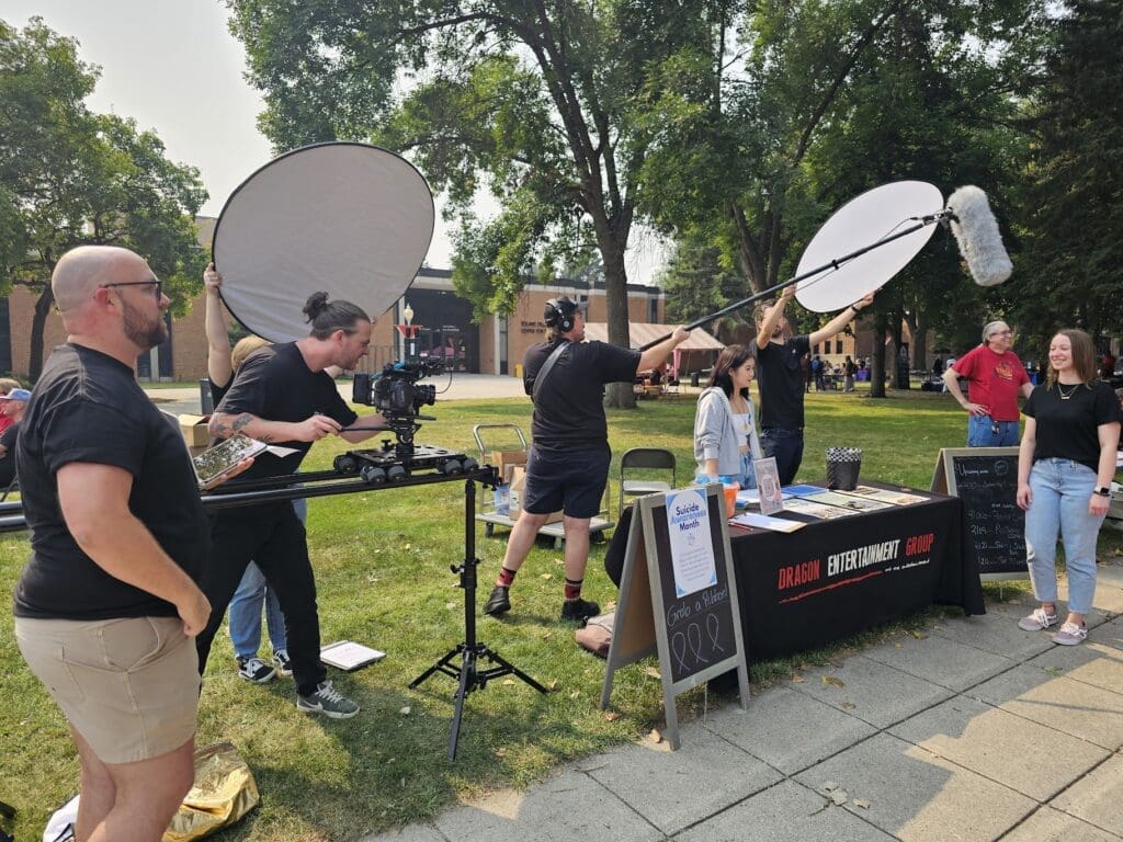 A film crew is outdoors, capturing a scene with a woman standing in front of a booth for 'Dragon Entertainment Group.' One crew member operates a camera on a slider, while another holds a boom microphone. Two additional crew members hold large reflectors to adjust the lighting. In the background, there are trees, buildings, and people attending what appears to be an outdoor event. A sign near the booth highlights Suicide Prevention Month.