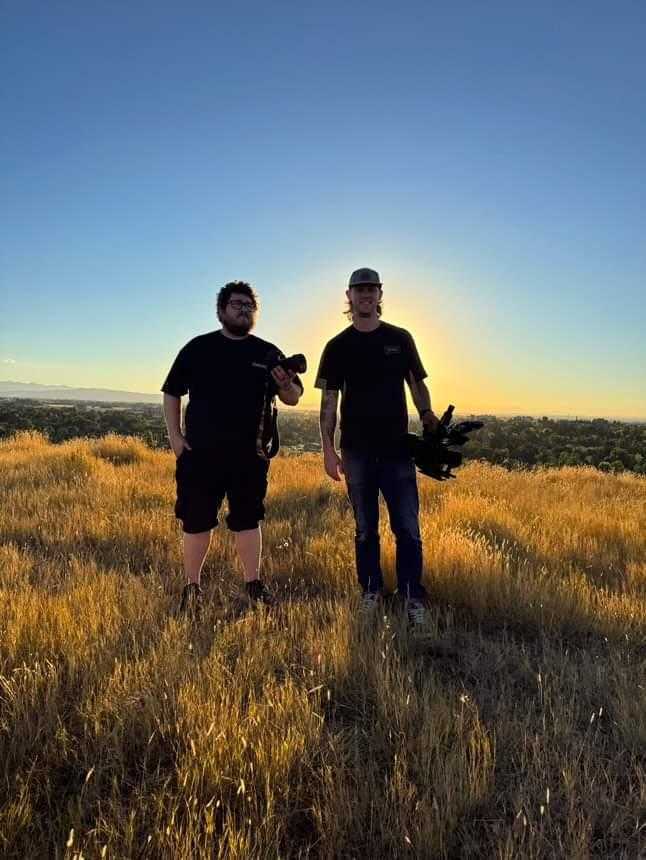 Two men standing in a grassy field, holding camera equipment, with the sun setting behind them, casting a golden glow. A valley with trees and distant mountains is visible in the background.