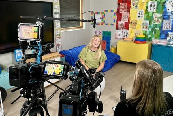 A behind-the-scenes view of a video interview in a colorful classroom. A woman sitting on a yoga ball is smiling while being filmed by two cameras on tripods. A 'Film Crew' member sits across from her, conducting the interview. The classroom is decorated with educational posters, colorful crafts, and soft furniture, creating a lively atmosphere.