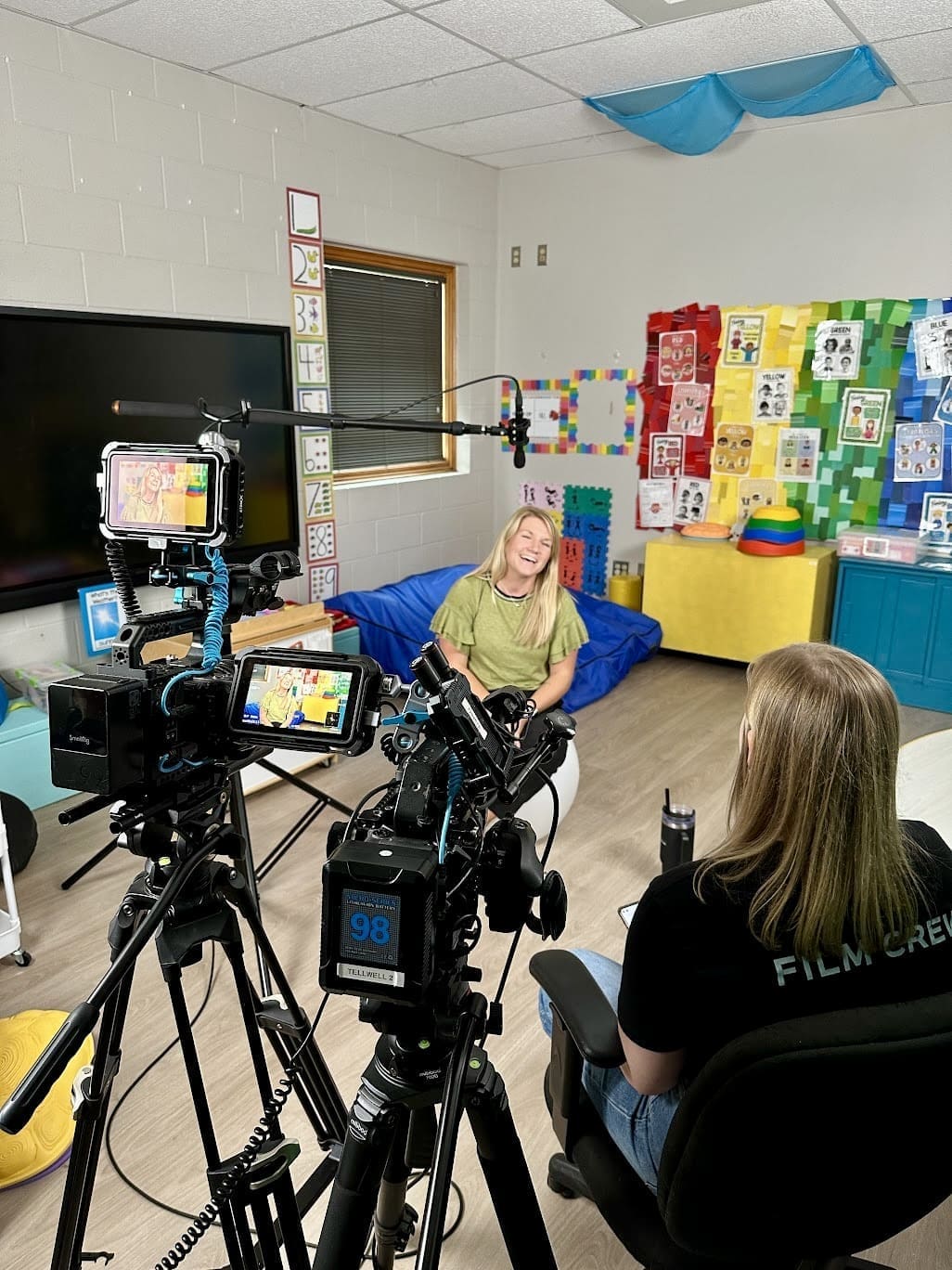 A behind-the-scenes view of a video interview in a colorful classroom. A woman sitting on a yoga ball is smiling while being filmed by two cameras on tripods. A 'Film Crew' member sits across from her, conducting the interview. The classroom is decorated with educational posters, colorful crafts, and soft furniture, creating a lively atmosphere.