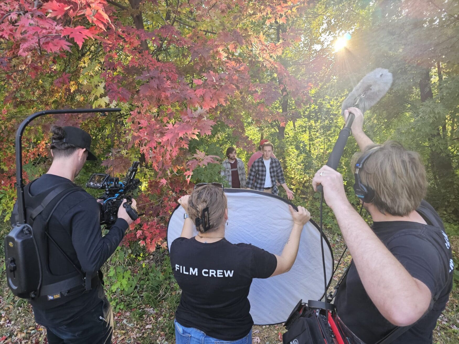 Outdoor image of trees with sunlight streaming through the fall green, red, and yellow leaves. Three film crew members are in the foreground facing away, towards three college-aged subjects walking towards the camera along a leaf strewn path.