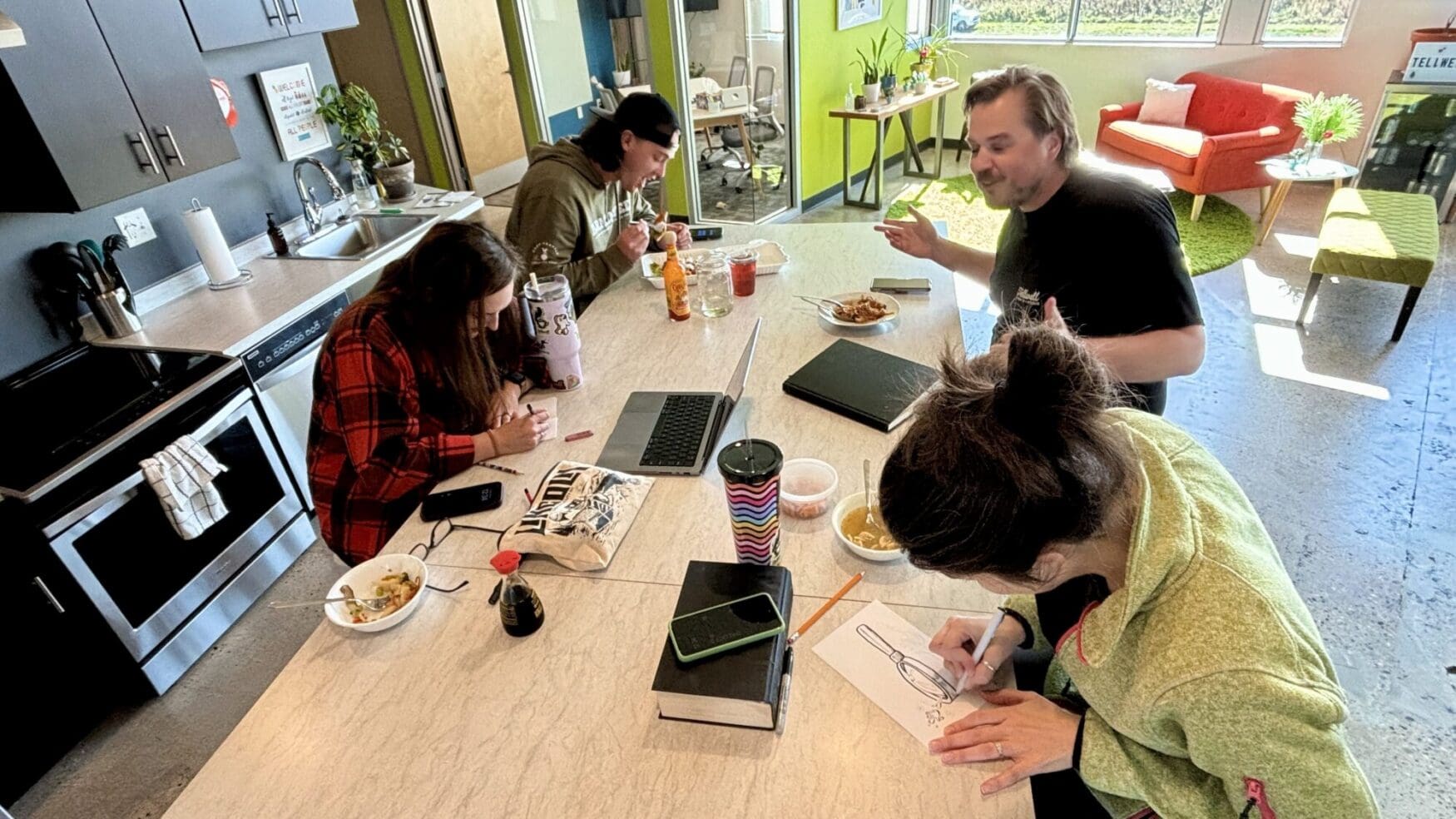 A group of four people sit around a kitchen island table in a brightly lit office, working on drawings, using a laptop, and eating lunch. The background includes a kitchen area and a sitting space with a red couch and large windows.