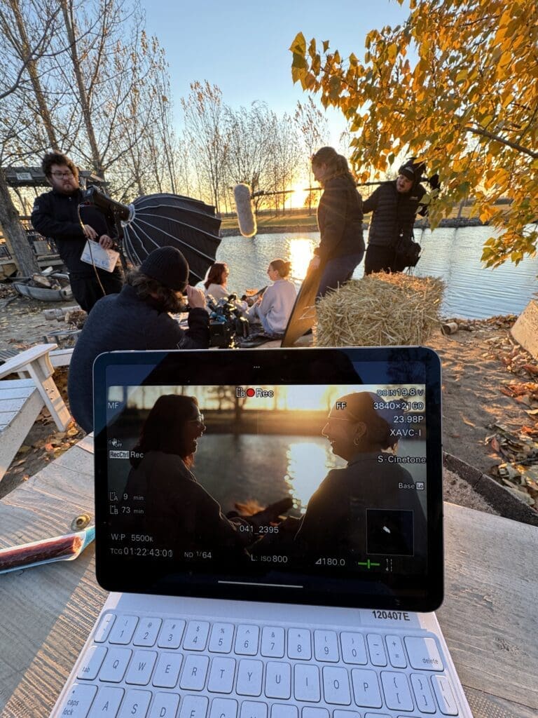 This image captures a film set at sunset by a lakeside. In the foreground, a tablet displays a live camera feed of two people in conversation with the sun setting behind them, casting a warm glow. Around them, a crew is setting up equipment, including a large reflector, a boom mic, and cameras. The autumn trees with orange leaves add to the cozy, natural ambiance of the scene.