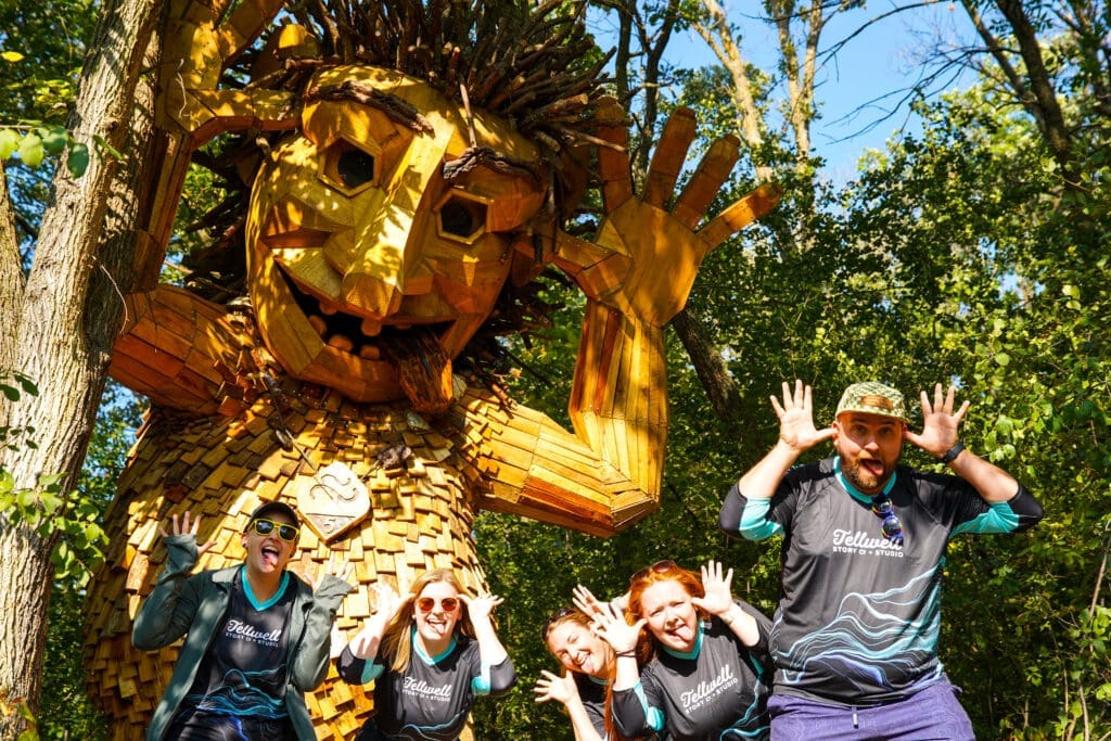 Group of people posing playfully in front of a large wooden troll sculpture in a forest. The group members are making funny faces and hand gestures, matching the troll’s playful expression. They are wearing Tellwell Story Co. + Studio shirts.