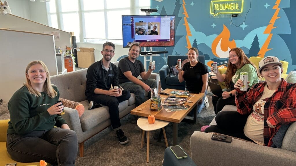 A group of seven coworkers sit around a coffee table in a casual office lounge area. They are smiling and raising cans in a toast while sitting on cozy couches, with a TV screen showing a virtual team member in the background.