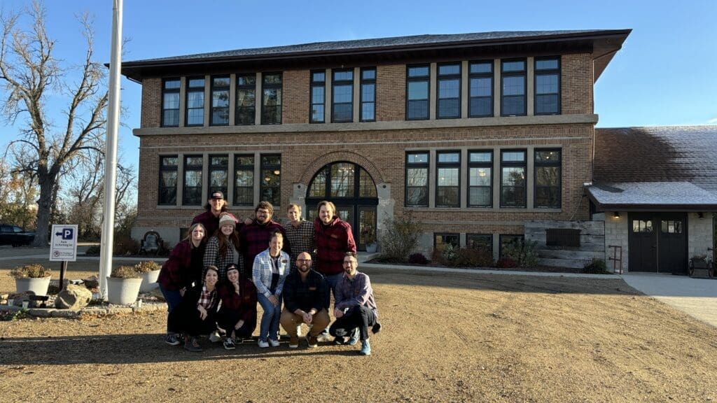 Group photo of a team posing in front of a large brick building with tall windows, under a clear blue sky. Everyone is smiling and wearing plaid shirts, standing or crouching together on a gravel path next to some large planter pots and a parking sign.