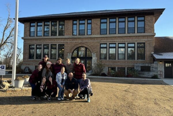 Group photo of a team posing in front of a large brick building with tall windows, under a clear blue sky. Everyone is smiling and wearing plaid shirts, standing or crouching together on a gravel path next to some large planter pots and a parking sign.