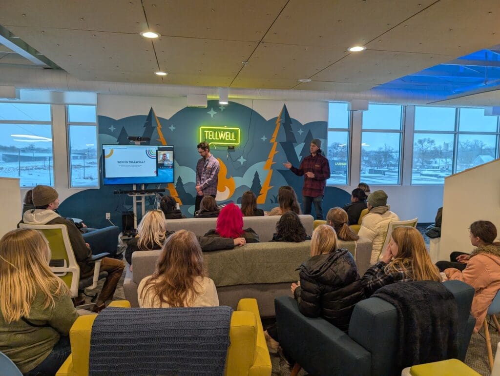 A group of about 10-20 college students sit together watching a presentation in a colorful office space. Two in person male presenters and one on a TV screen, in their late 20’s to early 30’s, stand in front of the students.