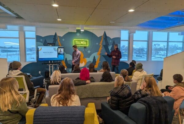 A group of about 10-20 college students sit together watching a presentation in a colorful office space. Two in person male presenters and one on a TV screen, in their late 20’s to early 30’s, stand in front of the students.