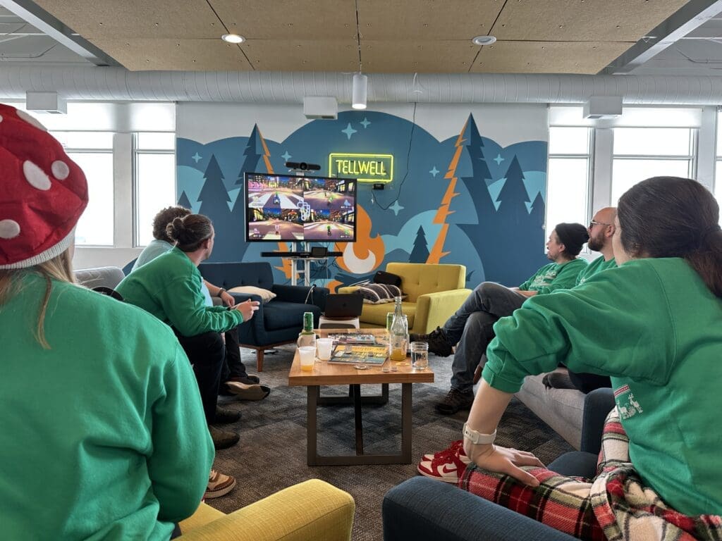 The image shows a room full of people wearing matching green sweaters with their backs to the camera while they play a video game on a large TV at the front of the room.