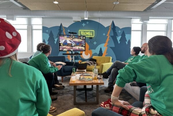 The image shows a room full of people wearing matching green sweaters with their backs to the camera while they play a video game on a large TV at the front of the room.