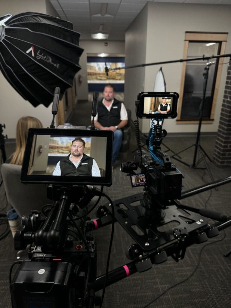 Video interview setup showing a seated man in a corporate hallway. Professional filming equipment, including cameras, monitors, a boom mic, and softbox lighting, is prominently displayed in the foreground.