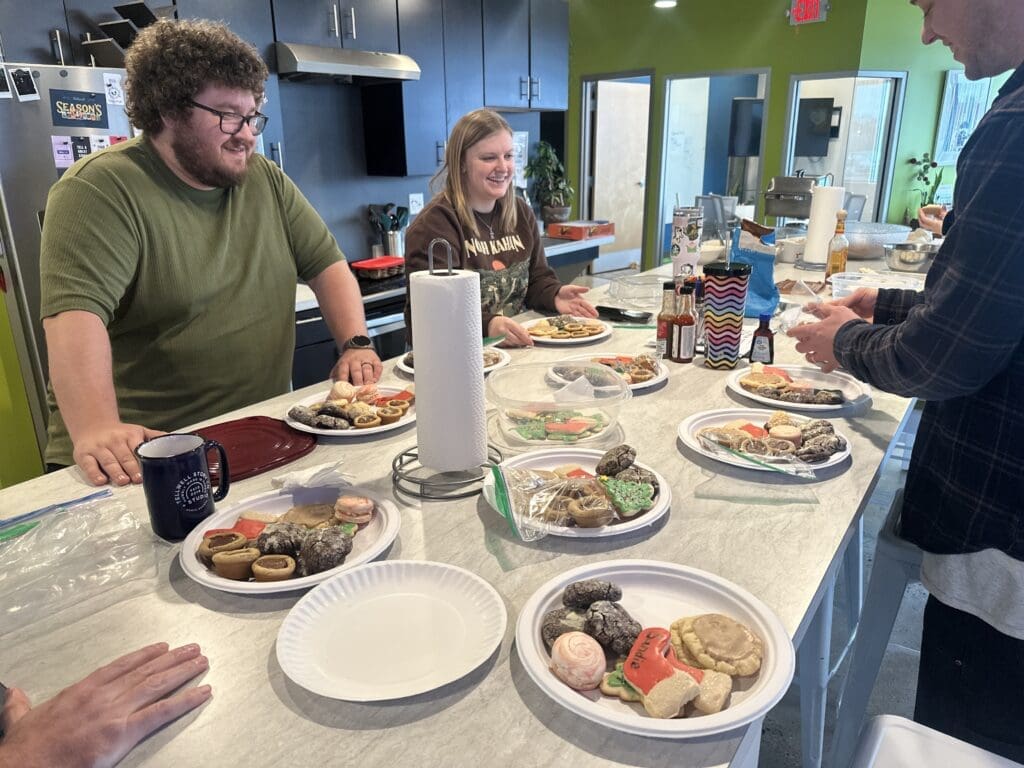 The image shows a group gathered around a kitchen island with plates of assorted holiday cookies. Two people, one in a green shirt and one in a brown sweatshirt, are smiling as they engage in conversation. Various cookie decorations and baking supplies are visible, creating a festive atmosphere in a modern kitchen setting.