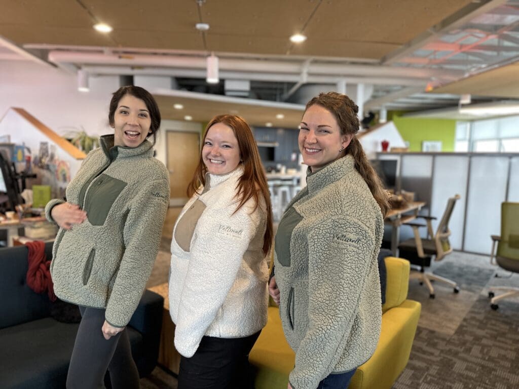 Three smiling people pose in a brightly lit office space, wearing matching sherpa jackets with the "Tellwell" logo embroidered on the sleeves. The office features modern furniture, including a teal couch and yellow chairs, with a background of desks and vibrant decor.