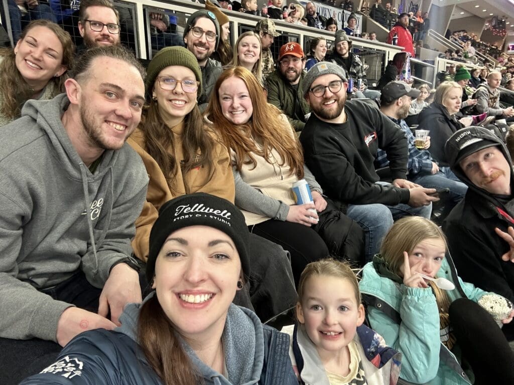 The image shows ​a lively group photo at a sports event, featuring adults and children sitting together in the stands, smiling and enjoying the atmosphere. The crowd is visible in the background, with some holding drinks and snacks. One person in the front is wearing a black beanie with the 'Tellwell Story Co. & Studio' logo.