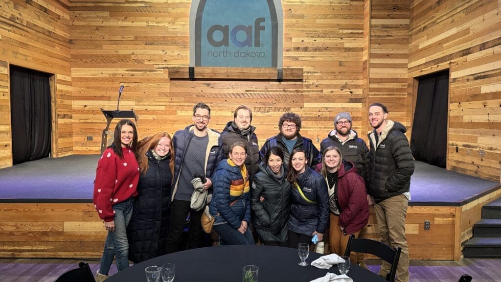 A group of smiling people posing in front of a stage with a wooden backdrop and a sign that reads "AAF North Dakota." The stage features a microphone and podium, and the group is dressed warmly in jackets, suggesting a winter event or gathering. Glasses and tableware are visible in the foreground on a table.