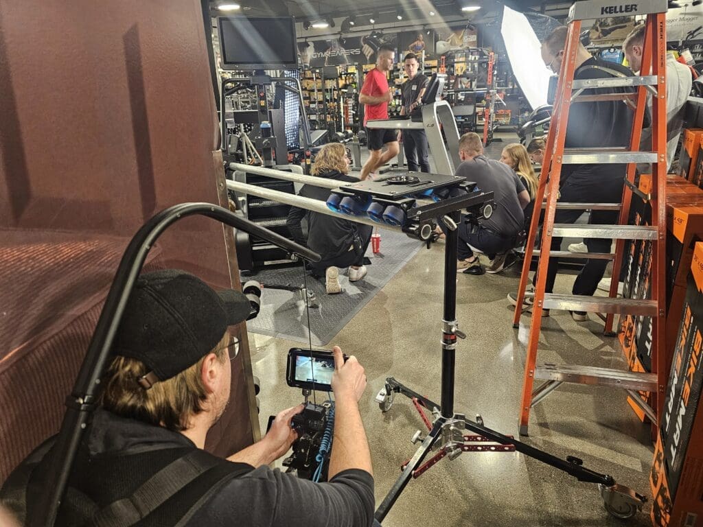 A behind-the-scenes look at a video production in a gym equipment store. A cameraman in a black cap and glasses films the scene with a handheld camera, while a group of crew members adjusts equipment near a treadmill where two men are engaged in a workout. A large light source illuminates the space, and a tall orange ladder stands nearby. The background is filled with gym machines, weights, and fitness-related products, creating a busy and professional filming environment.