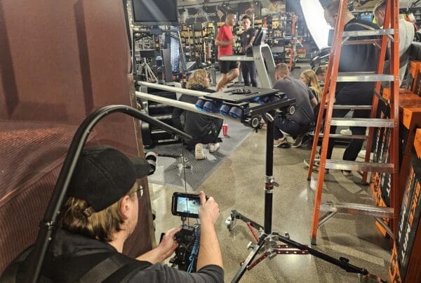 A behind-the-scenes look at a video production in a gym equipment store. A cameraman in a black cap and glasses films the scene with a handheld camera, while a group of crew members adjusts equipment near a treadmill where two men are engaged in a workout. A large light source illuminates the space, and a tall orange ladder stands nearby. The background is filled with gym machines, weights, and fitness-related products, creating a busy and professional filming environment.