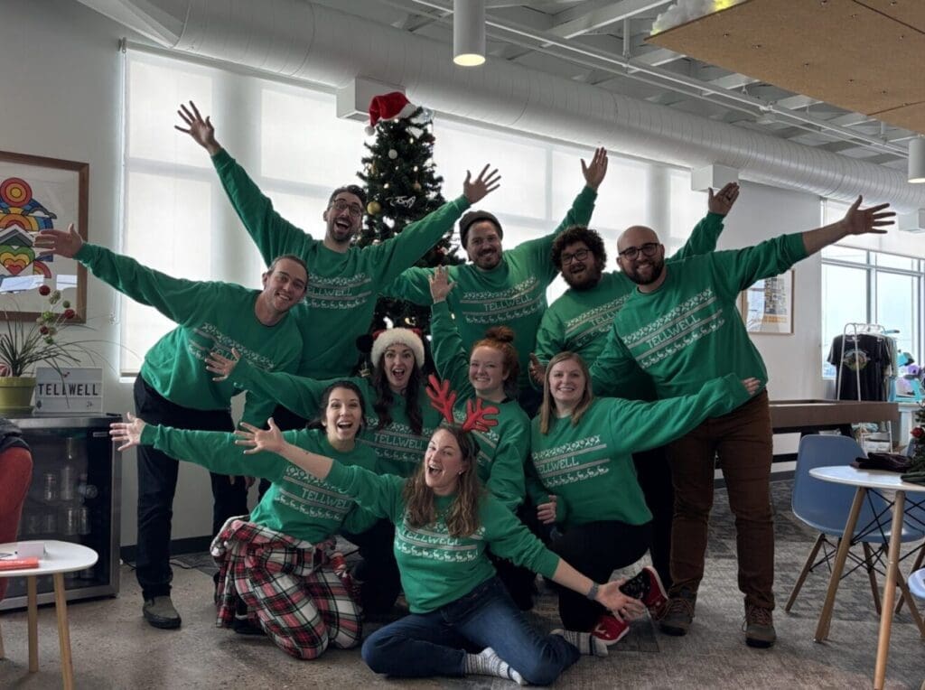 A group of people in matching green sweaters pose joyfully in an office with a decorated Christmas tree and festive decorations.