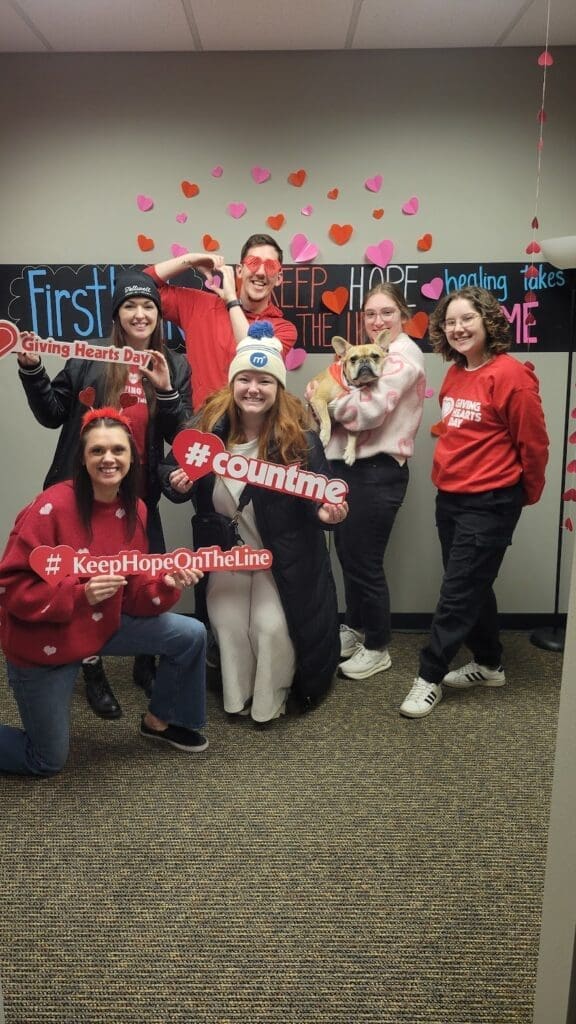 A group of six people is posing together in a decorated room for Giving Hearts Day. They are all dressed in festive red and black outfits, with some wearing heart-themed accessories like heart-shaped sunglasses and headbands. The background features a blackboard-style sign with colorful handwritten text that reads "Keep Hope On The Line" along with heart decorations in pink, red, and orange. Each person is holding a sign with messages related to the event, such as "#countme," "#KeepHopeOnTheLine," and "Giving Hearts Day." One person in the group is holding a small French Bulldog wearing an orange bandana. They are all smiling and striking playful poses, showing enthusiasm for the cause. The setting appears to be an office or community space with a carpeted floor and a neutral-colored wall.