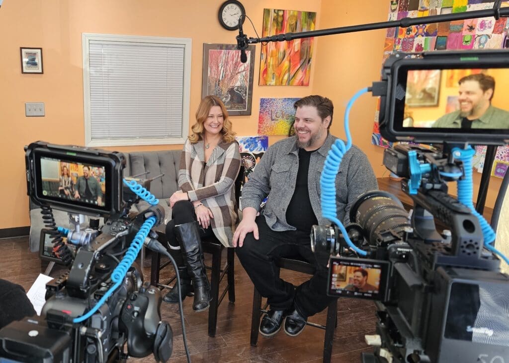 A behind-the-scenes view of a video interview setup in a colorful and artistic indoor space. A man and a woman sit on stools, smiling and engaged in conversation while looking at the interviewer or camera. The woman wears a plaid cardigan and knee-high boots, while the man wears a gray jacket over a black shirt. Two professional video cameras with blue cables and external monitors capture the scene, and a boom microphone is positioned above them. The walls are decorated with vibrant paintings, and a gray couch is visible in the background.