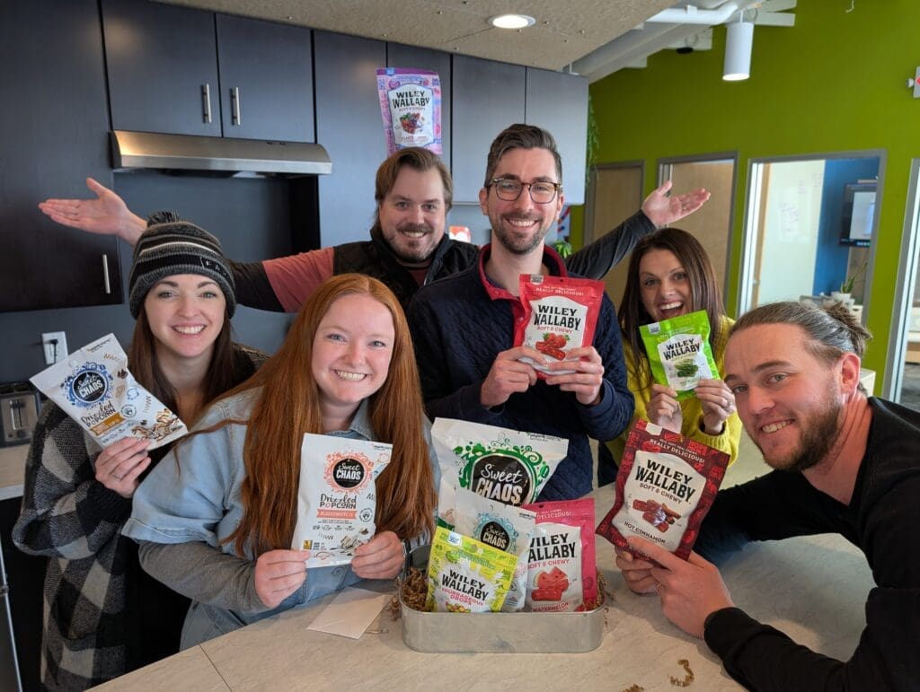 A group of six people gathers in a modern kitchen space, smiling and holding various snack products, including popcorn and Wiley Wallaby licorice. The background features dark kitchen cabinets and a bright green wall. The group appears cheerful and engaged.