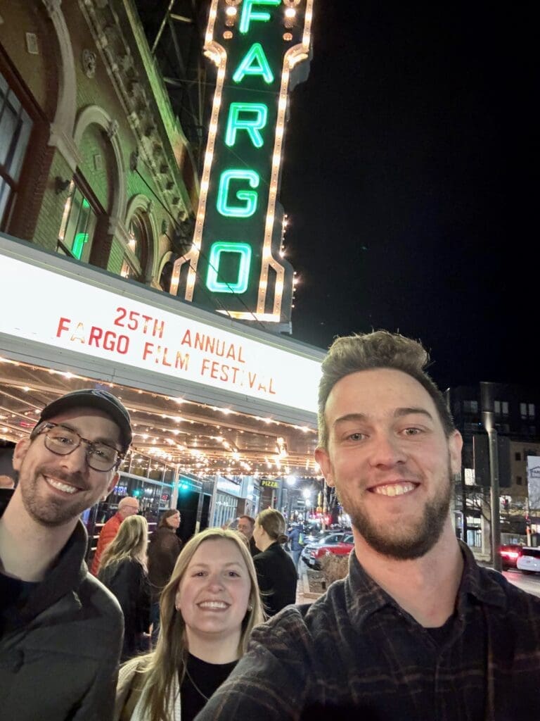 Three people smile for a selfie in front of the Fargo Theatre at night. The theater's iconic neon sign glows above them, and the marquee reads "25th Annual Fargo Film Festival." The person on the right, wearing a dark plaid shirt, holds the camera. The person on the left wears glasses, a black cap, and a dark jacket, while the person in the middle has blonde hair and is wearing a white sweater. The background is bustling with festival attendees and city lights.