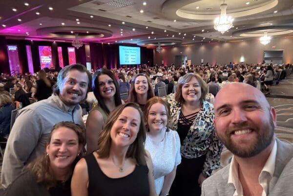 A group of eight smiling adults poses for a selfie at a formal banquet event in a large, elegant ballroom. The background shows a packed room full of people seated at round tables, with chandeliers overhead and colorful stage lighting on the far side reading “YWCA Women of the Year.” Everyone in the group looks happy and dressed in semi-formal to business casual attire.