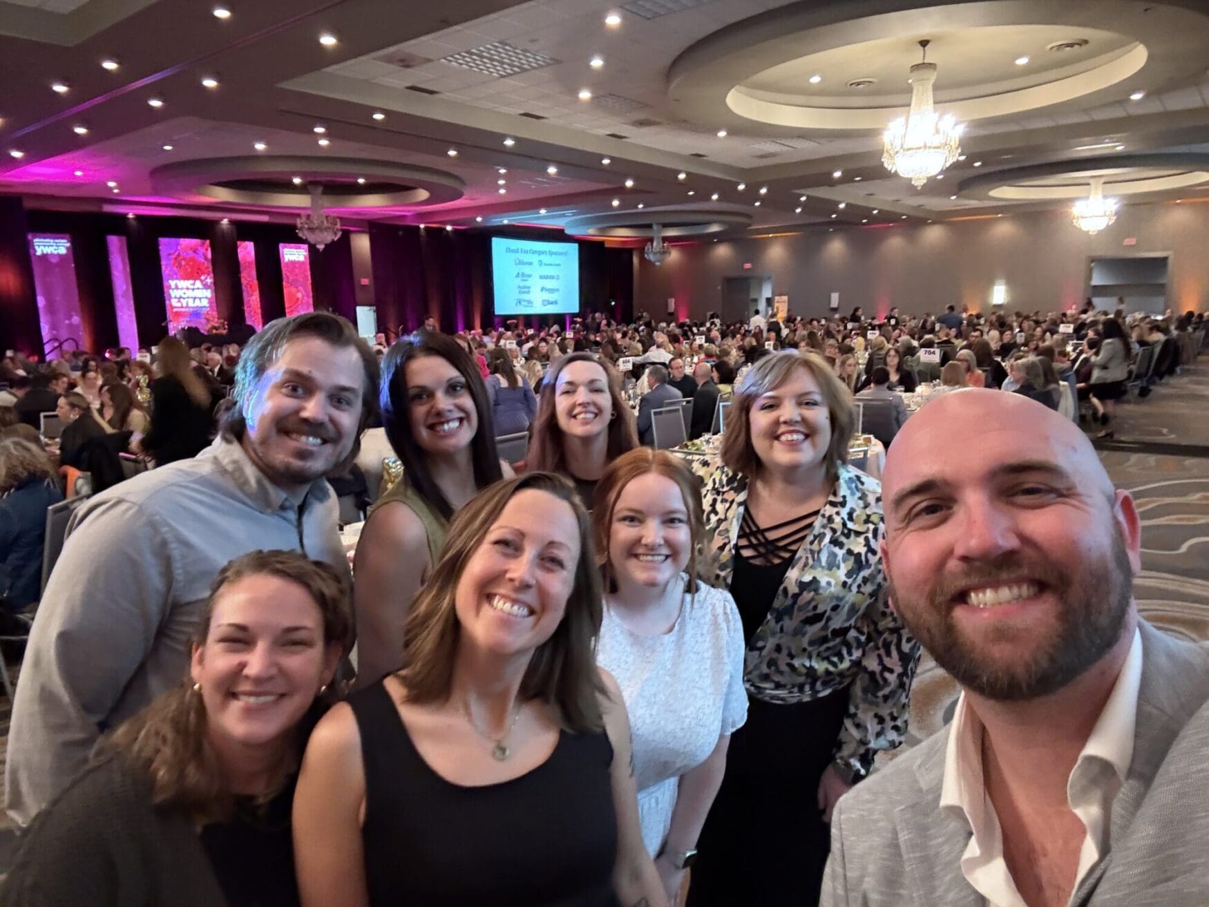 A group of eight smiling adults poses for a selfie at a formal banquet event in a large, elegant ballroom. The background shows a packed room full of people seated at round tables, with chandeliers overhead and colorful stage lighting on the far side reading “YWCA Women of the Year.” Everyone in the group looks happy and dressed in semi-formal to business casual attire.