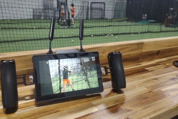 A wireless monitor on a wooden bench displays live footage of a young baseball player at bat inside an indoor training facility, while the practice continues in the background beyond a protective net.
