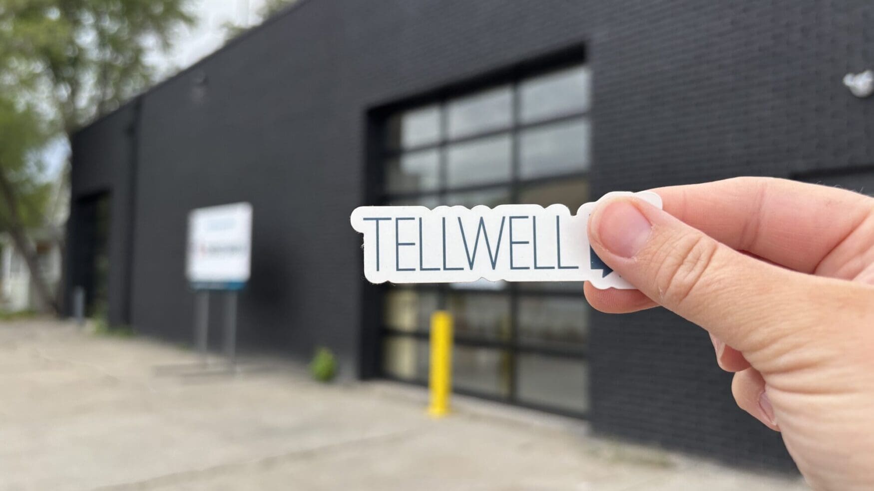 A hand holds a sticker that reads “TELLWELL” in front of a modern, black brick building with large glass garage-style doors. The background is slightly out of focus but includes a sidewalk, a standing sign, and some trees, suggesting the exterior of a creative or media office space. The image conveys a sense of branding or a new location reveal.