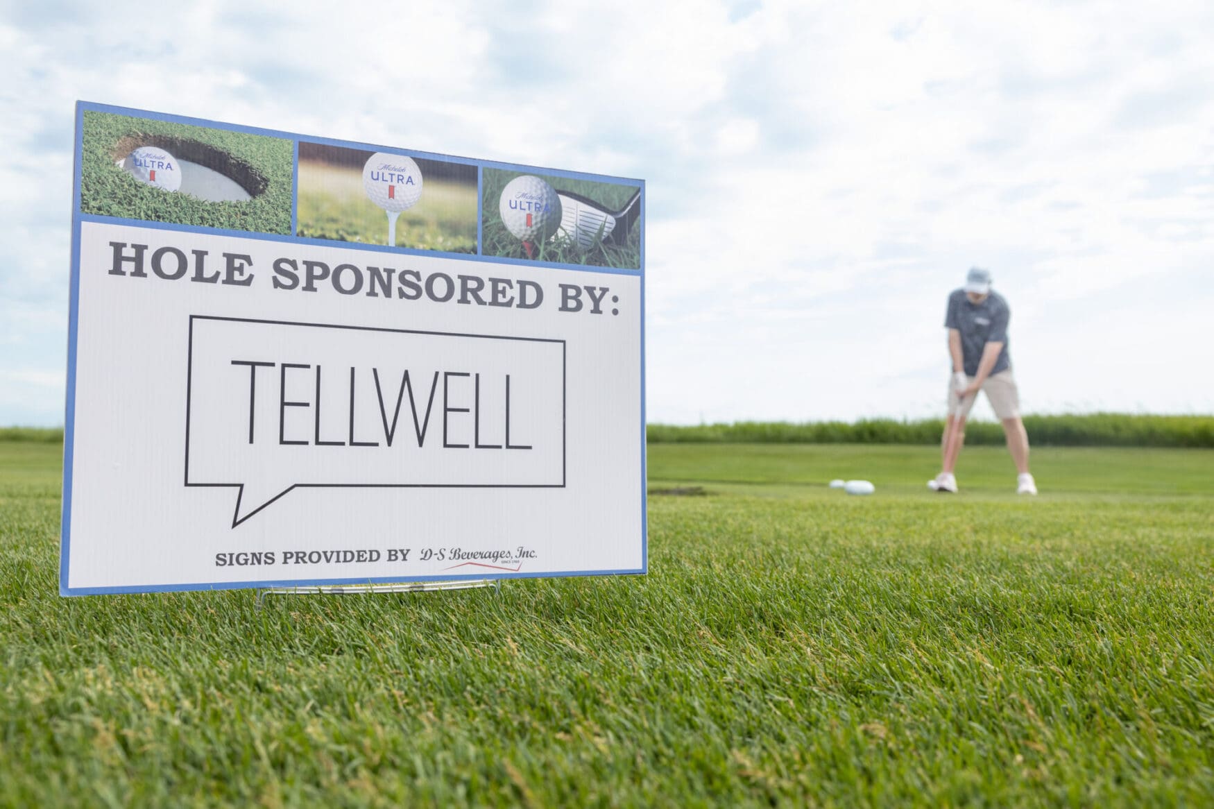 A sign reads "Hole sponsored by: Tellwell" is staked into the green of a gold course. There is a man preparing to swing a golf club in the background.