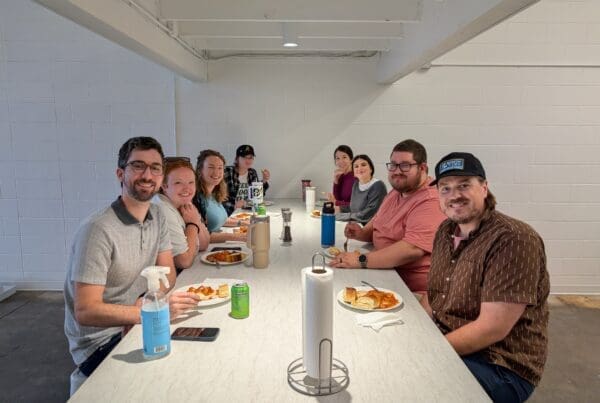 A group of 8 men and women sit at a large table sharing a meal. There are plates with pasta and garlic bread, an array of drink cans, and cleaning supplies on the table as well.