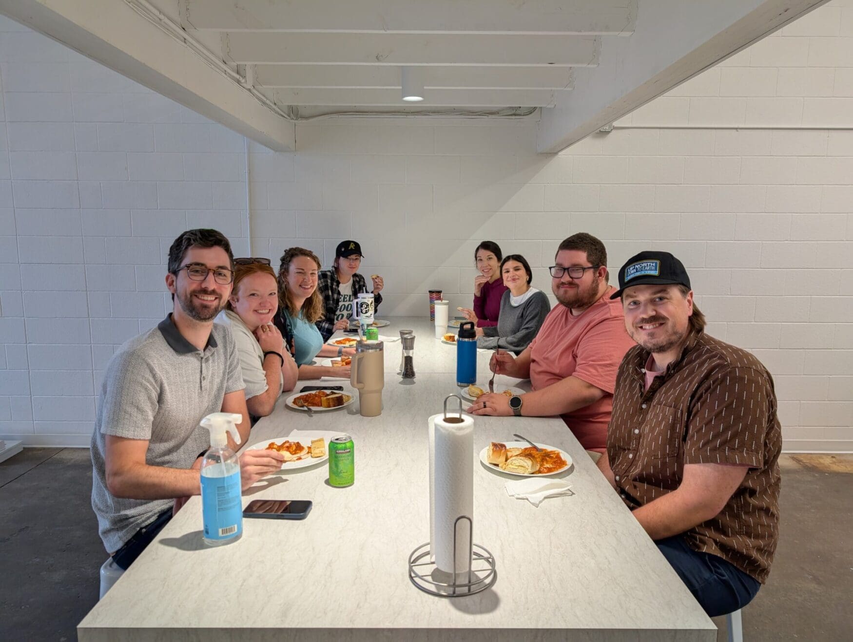 A group of 8 men and women sit at a large table sharing a meal. There are plates with pasta and garlic bread, an array of drink cans, and cleaning supplies on the table as well.