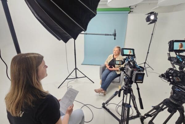 Behind-the-scenes of a video shoot in a white studio with a blue backdrop. A woman sits in a gray chair speaking on camera while another person off-camera holds interview notes. Multiple cameras, lights, and a boom mic are set up around her.