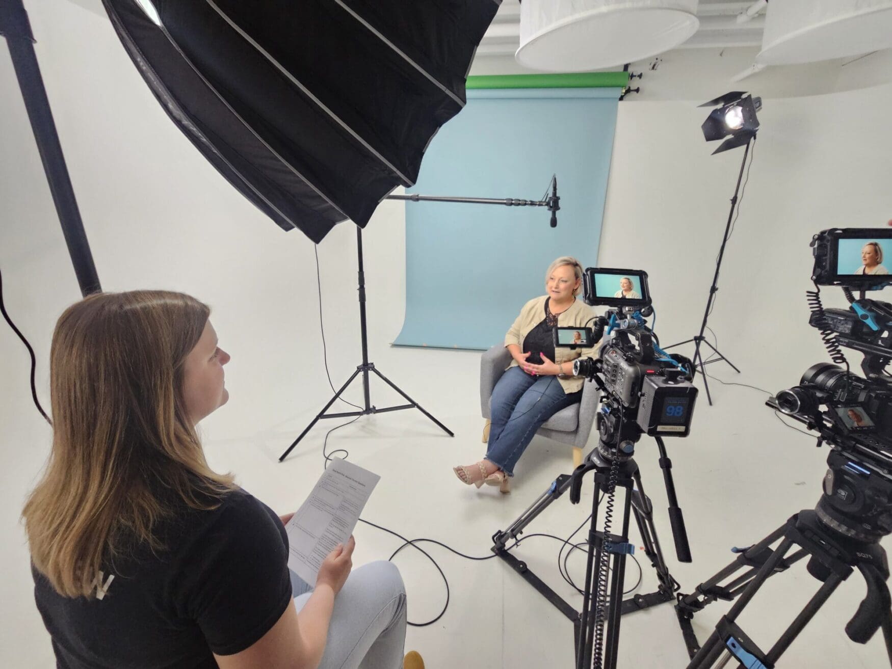 Behind-the-scenes of a video shoot in a white studio with a blue backdrop. A woman sits in a gray chair speaking on camera while another person off-camera holds interview notes. Multiple cameras, lights, and a boom mic are set up around her.