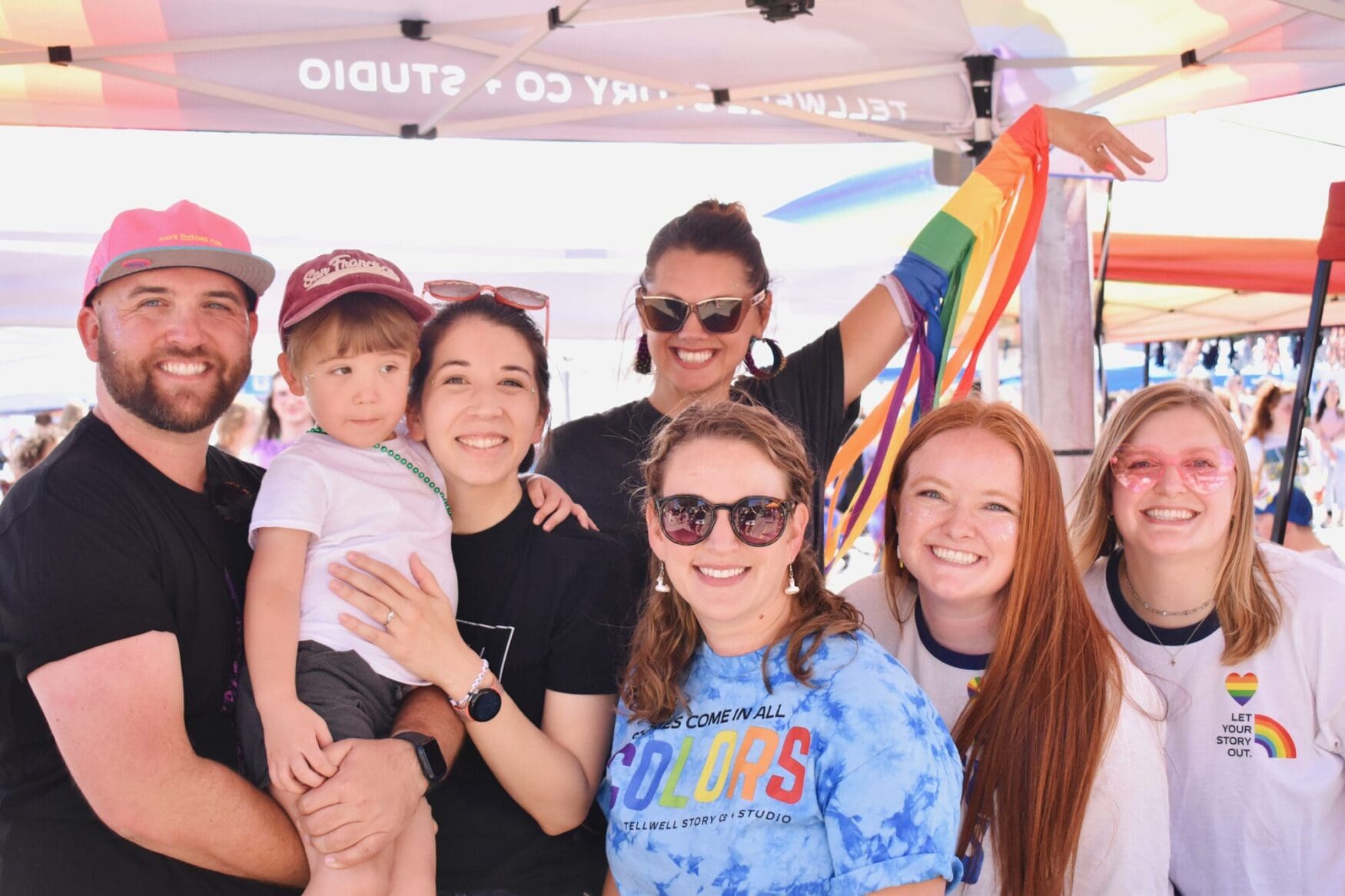 A group of six people, including a young boy, smile under a Tellwell Story Co. tent at a Pride event. Several are wearing rainbow-themed shirts, one person holds a rainbow flag, and everyone looks cheerful and celebratory.
