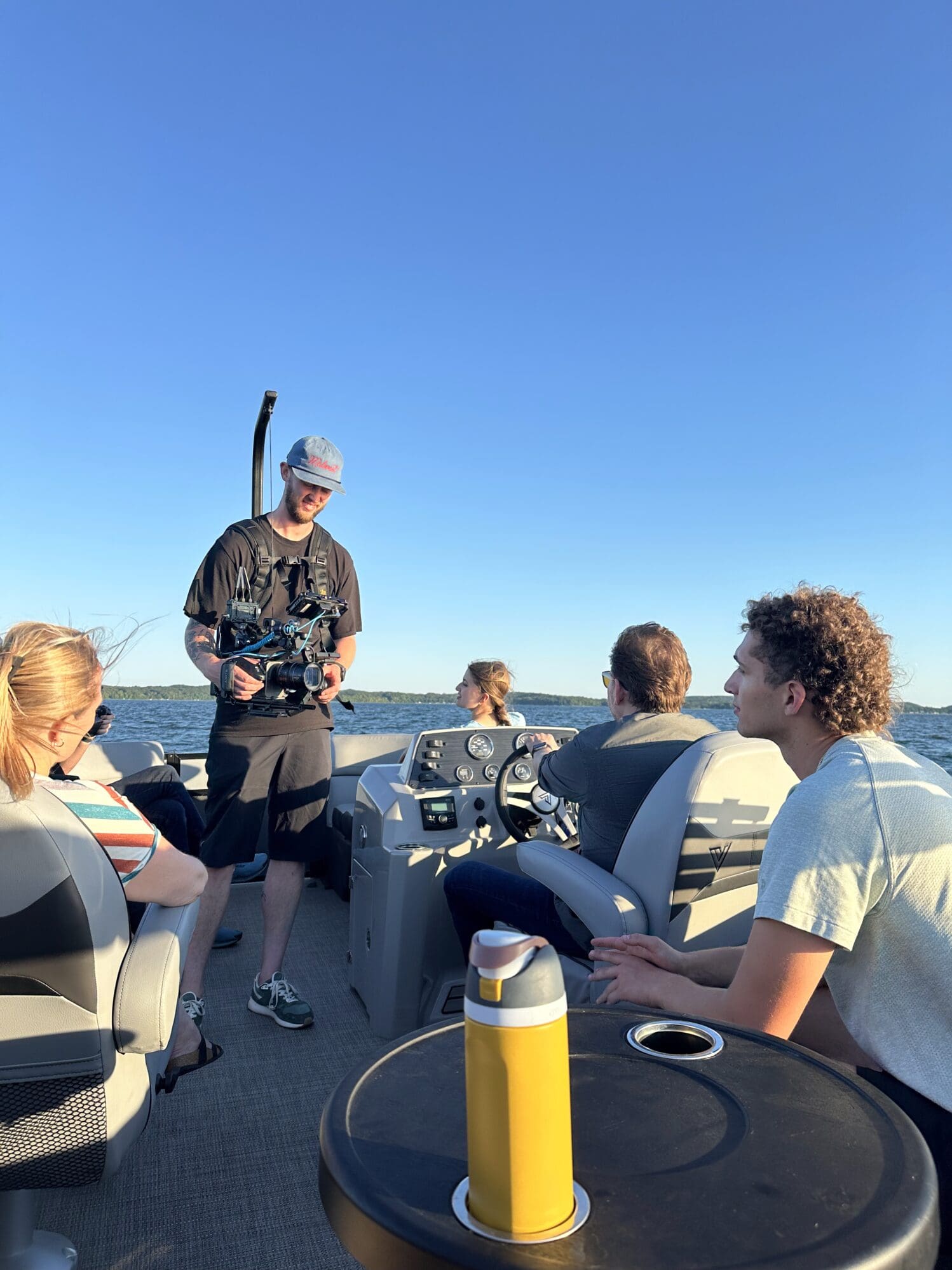 A group of people on a boat under a clear blue sky. One person is steering while others sit and talk. A cameraman with gear strapped to his chest is filming on deck. A yellow water bottle sits on a small table in the foreground.